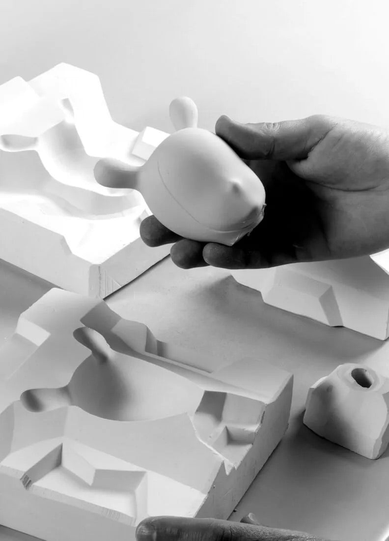 Hand holding a small white ceramic sculpture over a larger abstract white sculpture on a light gray background