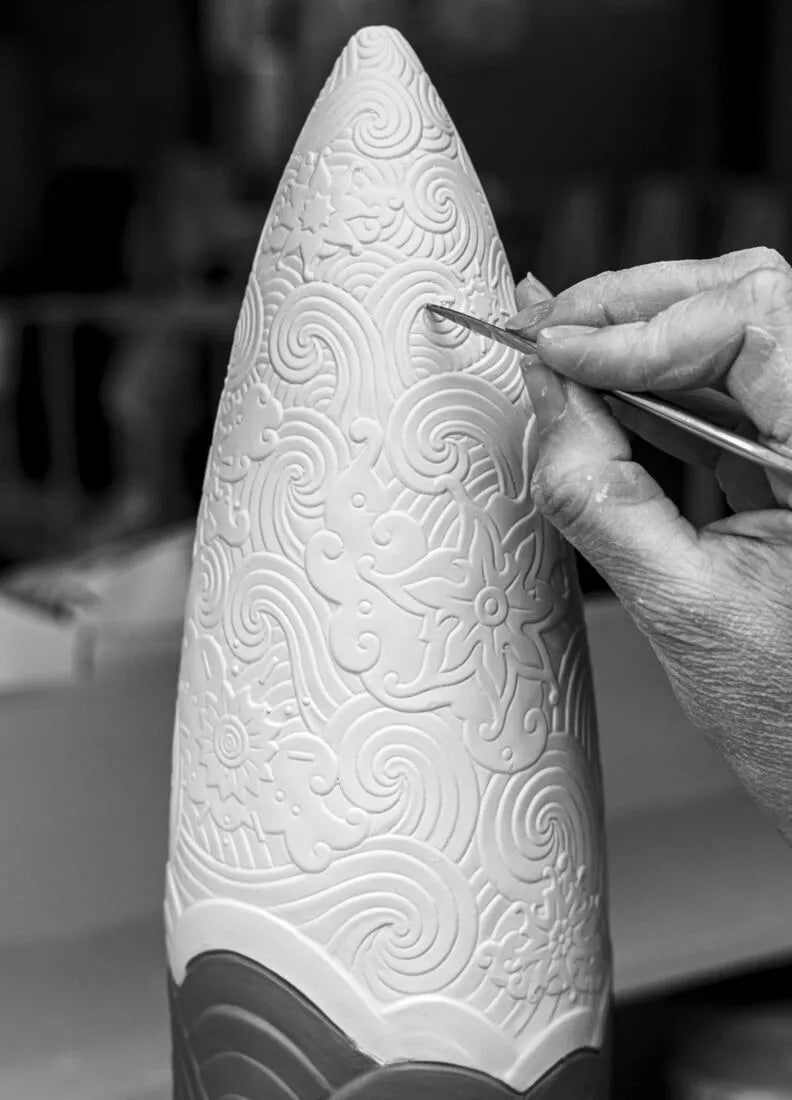 Person carving intricate patterns into a white cone-shaped object with a tool.