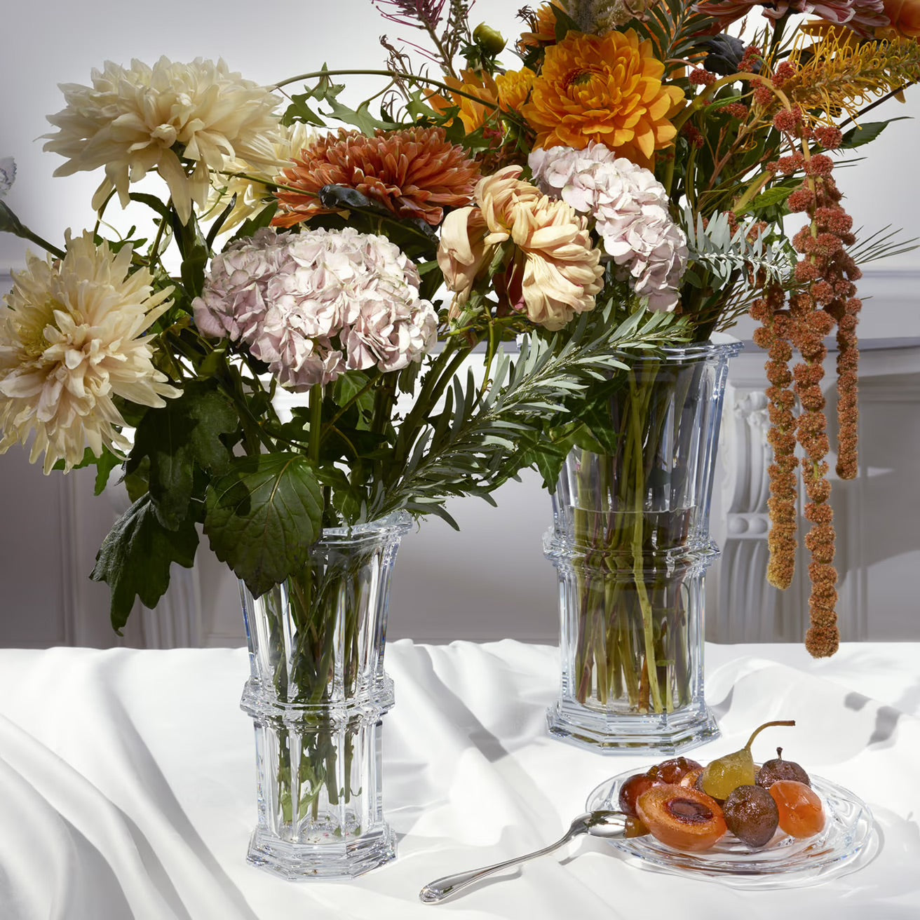 Three clear glass vases with flowers on a white tablecloth, with a small plate of fruits and a decorative item in the background.