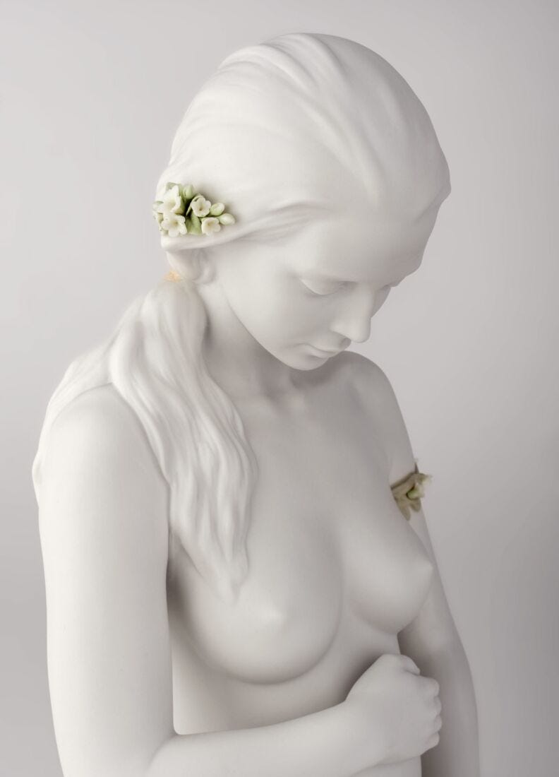 White classical-style female bust sculpture with long flowing hair, a small white floral hair accessory, and a small gold floral chest accent, photographed against a light gray studio background.