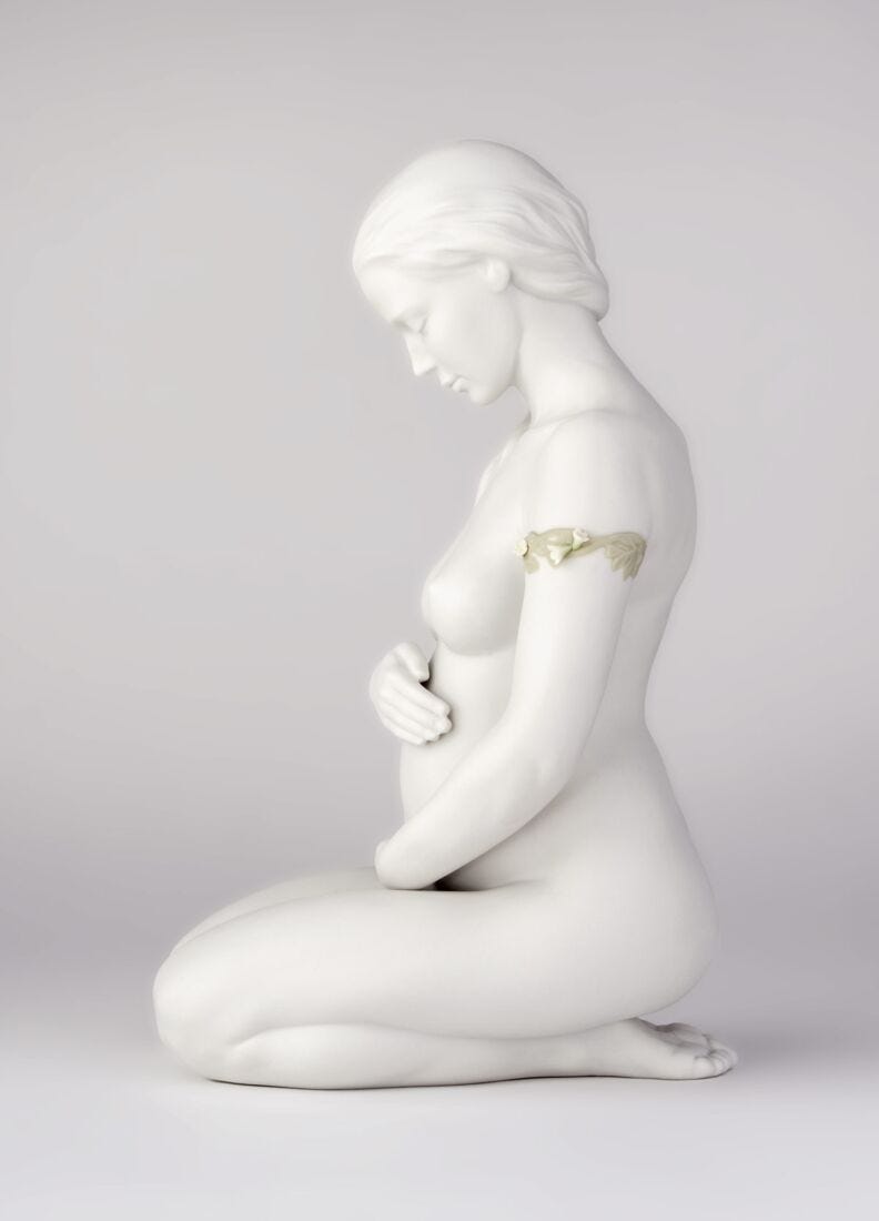 A white ceramic figurine of a kneeling pregnant woman in profile, featuring a delicate gold leaf detail on her upper arm, set against a neutral light gray background.