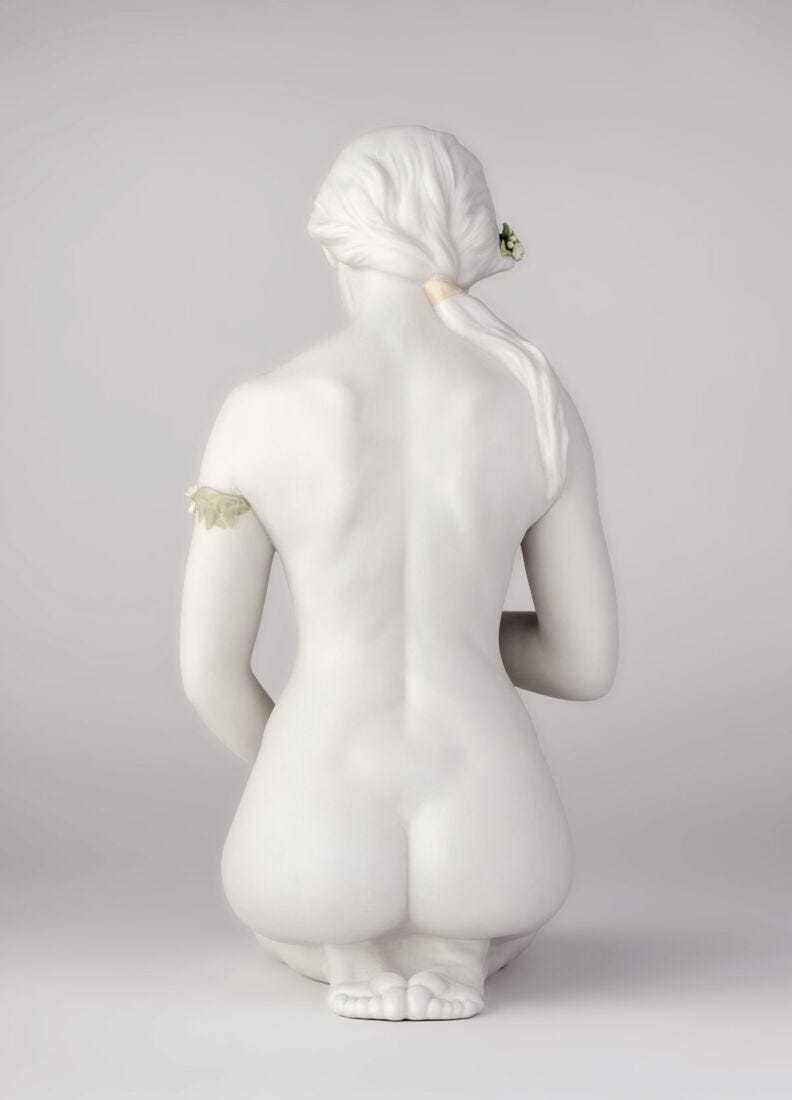 A white ceramic figurine of a kneeling nude female figure, shown from the back, featuring detailed sculpted hair, a small green flower in her hair, and a small leaf accent on her upper back, set against a neutral light gray background.