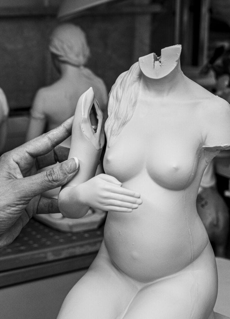 A black and white close-up of a sculpted female torso model with a missing head, being held by a hand in a workshop. Another blurred female figure is visible in the background.