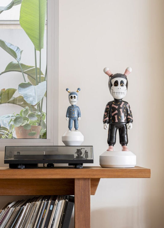 Two decorative skeleton figurines displayed on a wooden shelf in a home interior setting, with a window, potted plants, and vinyl records visible in the background.