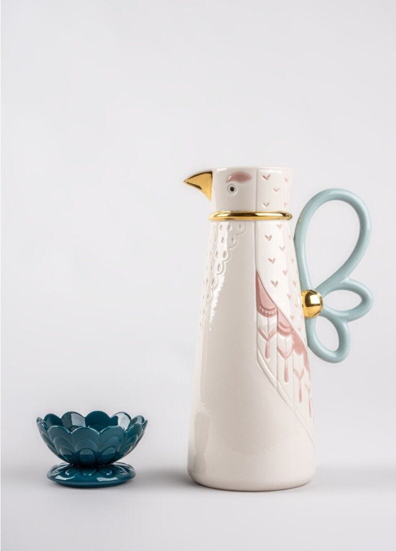 A ceramic bird-shaped carafe with a pale pink feather pattern, gold accents, and a light blue handle, shown next to a matching light blue flower-shaped bowl on a light gray background.