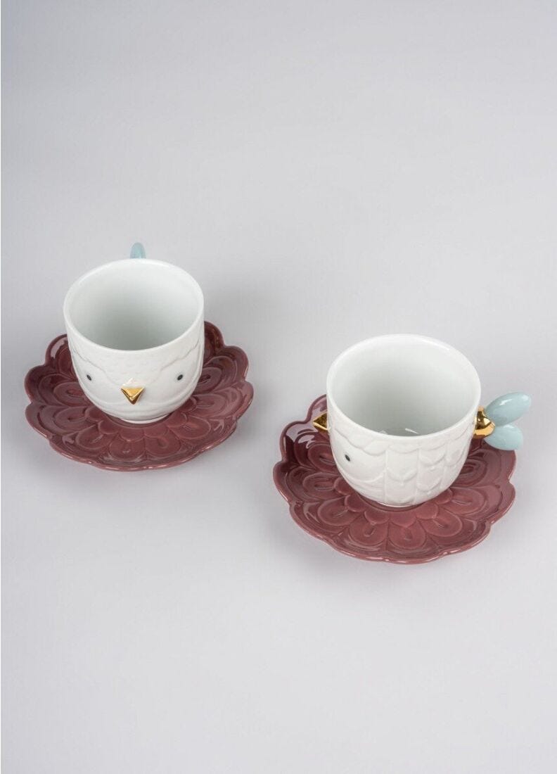 Two white ceramic owl-themed espresso cups with matching saucers, featuring a dusty rose patterned base, gold beak details, and light blue bird-ear handles, set against a neutral gray background.