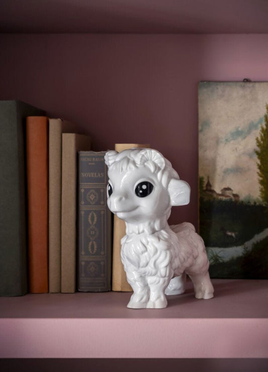 A white ceramic lamb figurine with large black eyes, displayed on a dusty pink bookshelf next to vintage books and a landscape painting.