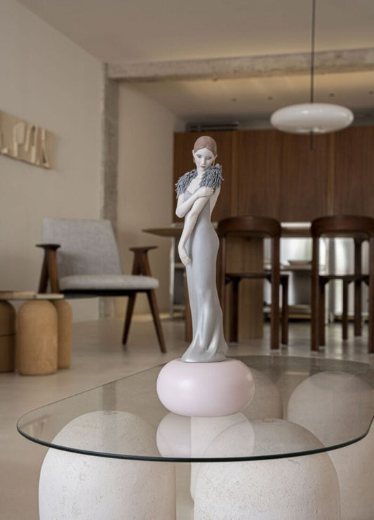 A white porcelain figurine of a woman with gray wings, standing on a pale pink round base, displayed on a glass-topped table in a modern, softly lit living space.