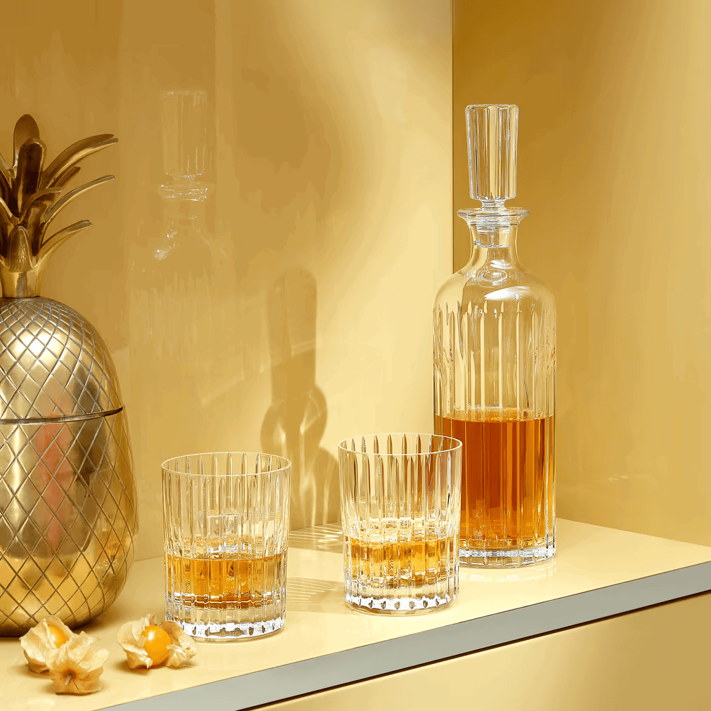 Two glasses and a decanter with amber liquid on a reflective surface with a gold pineapple and decorative elements.