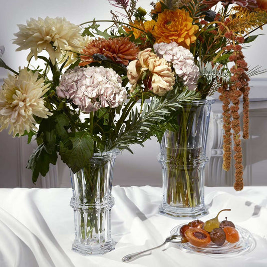 Three clear glass vases with flowers on a white tablecloth, with a small plate of fruits and a decorative item in the background.